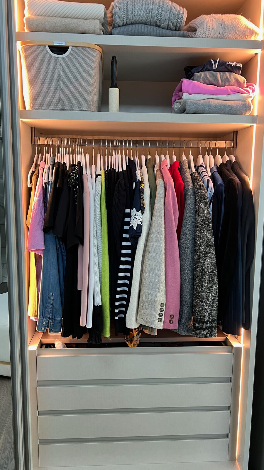 A lit fitted wardrobe with clothes hung by colour and jumpers folded on the shelves above