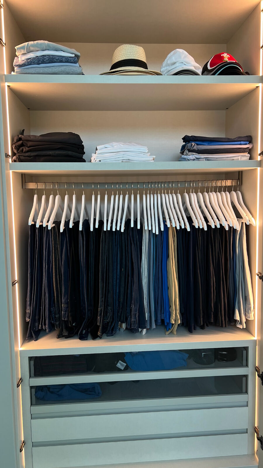 Trousers and jeans hung on matching white hangers, sorted from dark to light, with folded tops and hats above