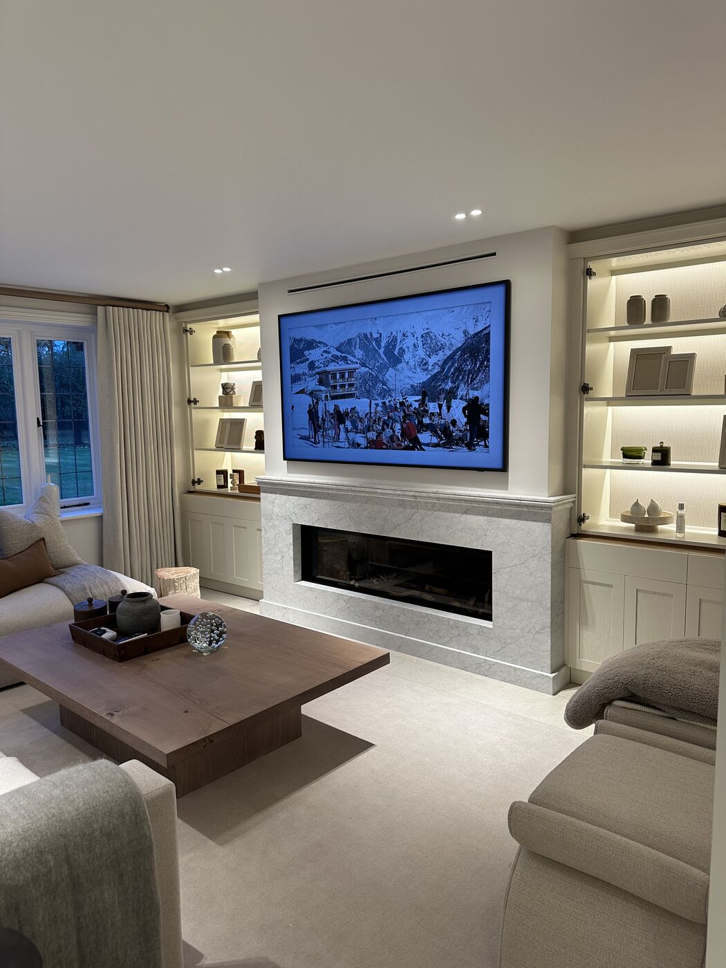 A calm, finished living room with lit shelving, a marble fireplace and soft neutral sofas