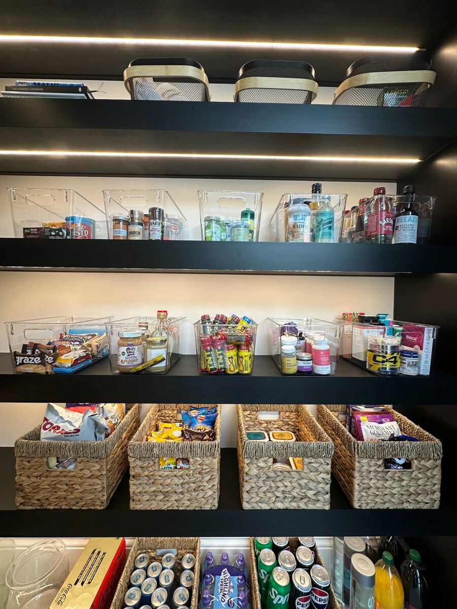 A lit walk-in pantry organised into clear tubs and woven baskets, with snacks, jars and drinks grouped by shelf